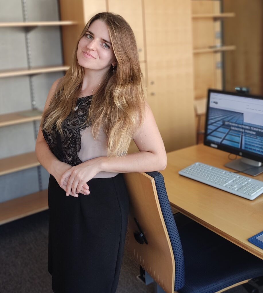 Marharyta Tsiura stands in front of a desk with a computer at the library, with her arms crossed in front, her head tilted towards the camera. She wears a sleeveless cream and black dress, and has long blonde hair.